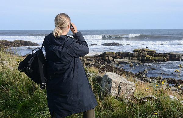Penny Martin stands in a grassy area by the seafront, looking our to the horizon. She wears a blue coat and a black backpack slung onto her left shoulder.
