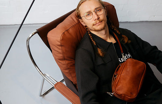 A man with light skin and brown hair in two plaits, wearing thin framed glasses, looks up at the camera from his seat in a arm chair. He has a black top on and wears a brown cross body bag.