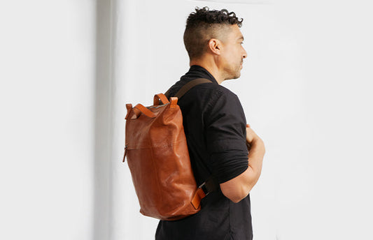 A man with tan skin and black hair, with shaved back and sides, stands facing away from the camera in a white room. He wears a black shirt and a large brown backpack on his back.