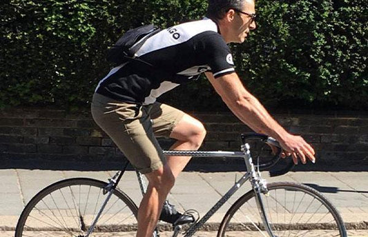 A man wearing a black and white t shirt and long shorts rides a bike on a road on a sunny day. There’s a hedge behind him and he wears a small black bag on his back.