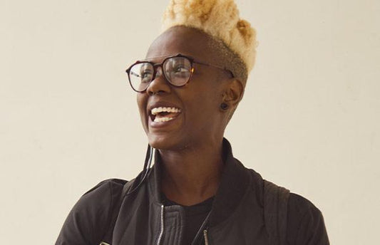 Natasher Beecher, with shaved back and sides and bleached blonde hair, smiles widely looking off into the distance. She wears tortoiseshell glasses and a black jacket.