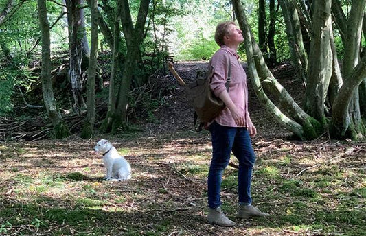 Sebastian Cox, in a shirt and jeans, stands, looking up in a grassy patch with trees in the background. He wears a backpack slung onto one shoulder. There is a small dog sitting to the left of him.