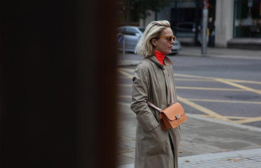 A woman with light skin and bleached blonde hair stands on the pavement with the road behind her. They are wearing an orange turtle neck, a beige coat, sunglasses and a small brown cross body bag.