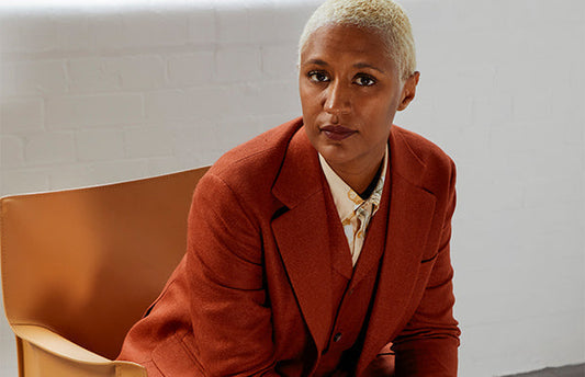 Maya Njie sits on a chair facing the camera. She wears a brick red suit and has a bleached blonde buzz cut